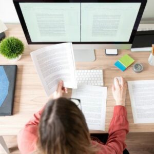 Woman sitting in front of a big desktop monitor with her back to us. She is comparing the text on a sheet of paper to the text in a word processor on the computer.